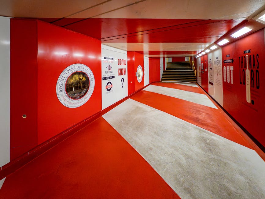 The image shows a brightly lit underground pedestrian walkway with red walls and ceiling, and a white and red patterned floor. On the left wall, there is a circular logo for Regent's Park, and to its right, informational posters and signs with questions such as 'Did you know?' displayed alongside a London Underground roundel symbol. The walkway has a set of stairs at the far end leading upward, and the corridor appears clean, modern, and designed for safe, efficient passage. This environment is suitable for a home relocation context, where furniture transport and packing may involve navigating such walkways during moving services. Man and Van Marylebone often orchestrates this type of logistics, ensuring furniture and boxes are carefully carried through accessible routes during house removals.