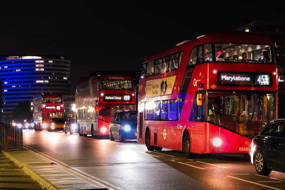 A nighttime scene featuring multiple red double-decker buses parked along a street in Marylebone, London, with the bus on the right displaying the route number 453 and destination 'Marylebone' in illuminated digital signage. The buses are lined up behind a dark-colored car, with the front bus showing a large advertisement on its side. Bright streetlights reflect off the bus windows, revealing passengers inside, and a nearby modern building with blue lighting is visible in the background. The scene captures a typical urban setting during evening hours, with street parking and traffic lanes marked on the pavement, illustrating the transport infrastructure supporting house removals and moving logistics in Marylebone, central London. Man and Van Marylebone occasionally operates in the area for home relocation, furniture transport, and packing and moving services, possibly utilizing such buses or nearby roads for moving logistics.