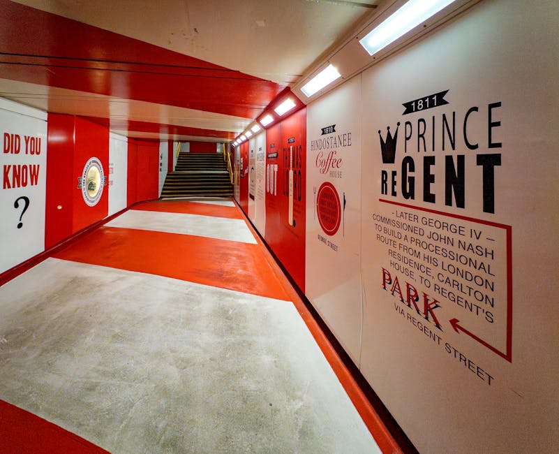 A signal box or underground station corridor with a curved, white concrete floor featuring a central red strip. The walls are painted with large, bold advertisements and informational posters in red, black, and white. On the right side, there is a prominent sign with the word 'PRINCE REGENT' and a crown graphic, along with historical details about George IV and local building developments. The corridor is illuminated by a series of fluorescent light fixtures on the ceiling. At the end of the corridor, a staircase with black and yellow striped safety markings on the steps leads upward. The environment is clean and well-lit, suggesting it may be part of a heritage site or transport-related setting. Occasionally, the corridor is used for context in house removals or moving services, such as transporting furniture or boxes through similar indoor spaces, with [COMPANY_NAME] providing logistical support for home relocation and furniture transport projects.