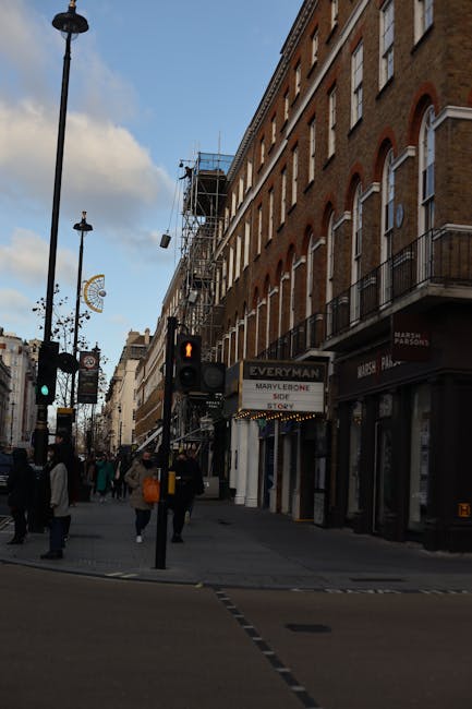 View of a busy Marylebone street during daytime, featuring a multi-storey brick building with several windows, some equipped with black metal balconies. The ground level shows storefronts, including a sign for Marple, a costume shop, and a partially visible shop front for Marie Parsons. A scaffolding structure is erected on the side of the building, indicating ongoing maintenance or renovation work. Pedestrians are walking along the pavement, some carrying shopping bags, while others pause at the traffic light, which is showing a red signal. A street lamp and traffic signs are also visible, with part of a classic London clock in the background. This urban scene reflects typical city activity relevant to household relocations, packing, and furniture transport logistics managed by companies like Man and Van Marylebone, especially for house removals or flat clearance services in Marylebone.