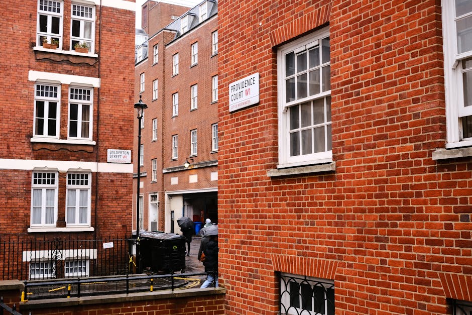 View from a high window overlooking a wet street in Marylebone during rainy weather, with pedestrians walking under umbrellas and crossing the road. The street is lined with red brick residential buildings with multiple bay windows and storefronts, including a pharmacy with a green cross sign. A vehicle is parked close to the curb, and a van is partially visible on the street. The sidewalk features a street lamp and various traffic signs, while the wet pavement reflects the overcast sky. This scene depicts an urban environment suitable for house removals and furniture transport, as part of a typical home relocation process, with Man and Van Marylebone likely involved in assisting with packing, loading, and transport logistics.