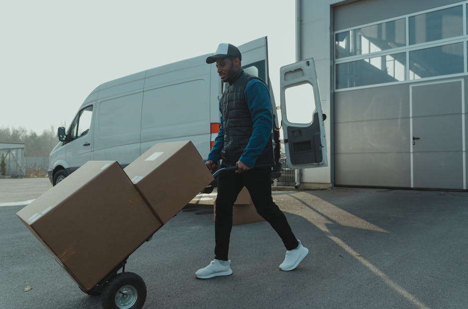 A man dressed in a blue jacket, black vest, black pants, and white sneakers is pushing a hand truck loaded with two large cardboard boxes outside a warehouse or industrial building. The loading area features a large, open vehicle door revealing a spacious interior with a visible van parked nearby, which is part of a professional removals and home relocation service by Man and Van Marylebone. The scene is set on a paved surface under natural daylight, with the van positioned adjacent to the building's exterior wall and some greenery visible in the background. The man appears to be engaged in the loading process, preparing to transport packed items as part of a furniture transport or packing and moving operation, with additional cardboard boxes lying on the ground nearby, indicating an active moving or packing session.