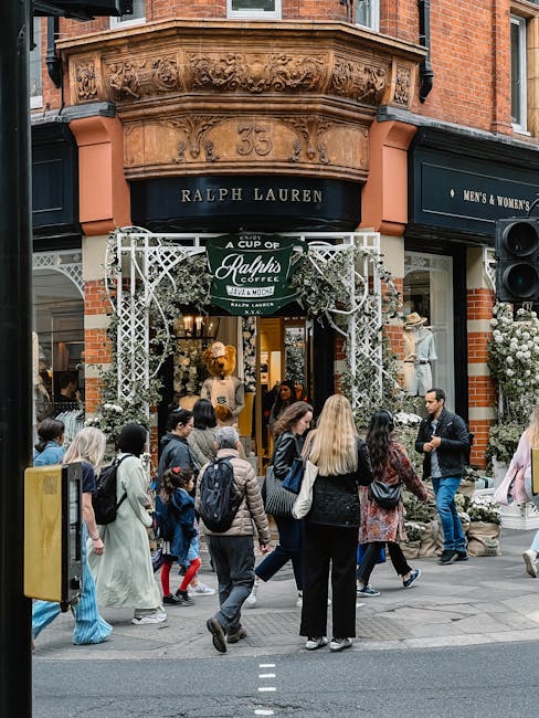 A busy street scene outside Ralph Lauren on Marylebone High Street, with multiple pedestrians walking and standing in front of the store. The storefront features a black sign with gold lettering, and an ornate, decorative archway made of white latticework and greenery, indicating a possible promotional event or outdoor display related to packing or moving services. Inside, through the open entrance, clothing displays are visible, including mannequins dressed in stylish attire. The street surface includes asphalt and paving stones, with pedestrians carrying backpacks and shopping bags, some wearing jackets and casual clothing. The surrounding buildings are constructed from red brick, with architectural details and additional signage for retail outlets, reflecting a typical boutique shopping area in Marylebone. The scene captures the movement and activity associated with house removals or furniture transport in a commercial environment, with [COMPANY_NAME] occasionally referenced naturally as part of the context.