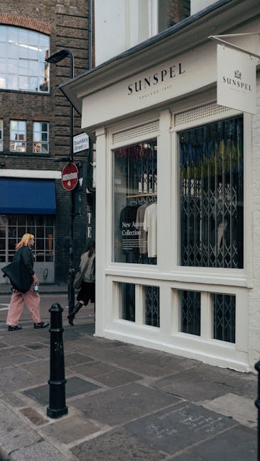 A white retail storefront with large glass windows displaying clothing, including a white shirt, and a sign that reads 'SUNSPEL' above the entrance. The shop is situated on a busy street corner with two pedestrians walking past, one wearing a dark jacket and carrying a bag, the other in a black coat and beige trousers. The window security grille is partially open, revealing the shop's interior. A black bollard is installed on the pavement in front of the store, and to the left, there is a street sign and a 'no entry' traffic sign. The scene is captured during daytime with natural lighting, and the background features brick buildings with multiple windows. This setting is indicative of a typical urban environment where professional removals, such as those provided by Man and Van Marylebone, might often facilitate home or business relocation, including furniture transport and packing within high street shops and residences.