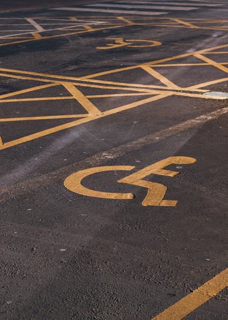 A close-up view of a dark asphalt parking lot surface showing multiple designated parking bays marked with yellow lines. Prominently visible in the foreground is a yellow-painted disabled parking symbol and a corresponding black and yellow striped barrier area. In the background, additional parking bays are marked with yellow overlapping lines and the number '2', indicating reserved or specific use spaces. The surface appears clean with some minor wear and tire marks. This parking lot setting is typical for house removals or furniture transport logistics, supporting loading and unloading processes used by [COMPANY_NAME] during home relocation arrangements in the Marylebone area. The image reflects the organized layout for vehicle access, permit compliance, and efficient loading for moving services.
