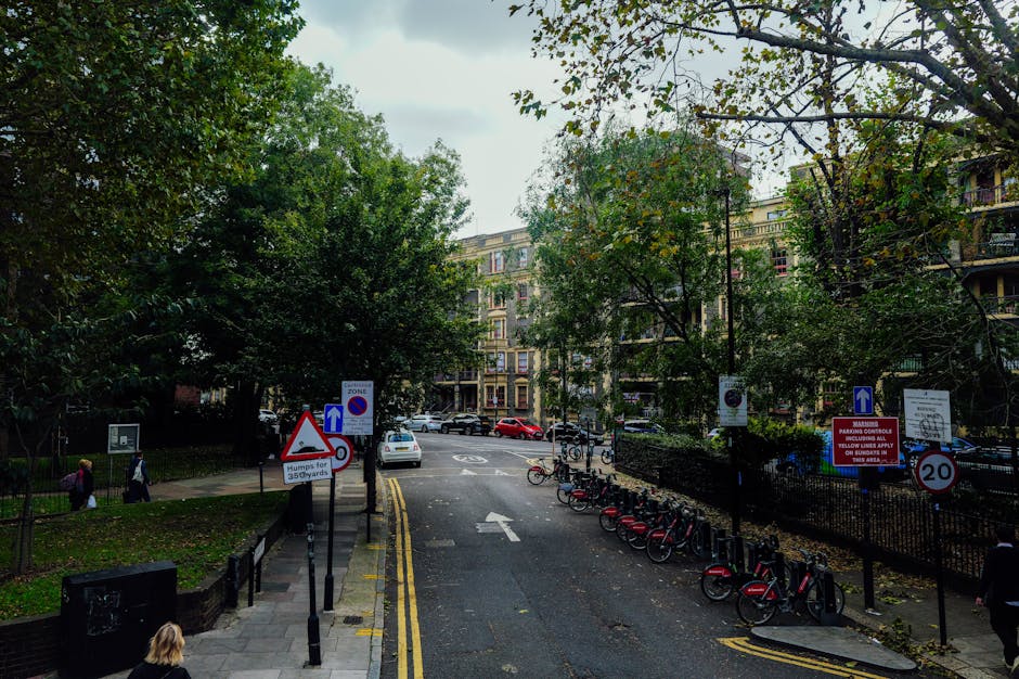 A street scene in Marylebone showing a narrow road with a row of parked bicycles on the right side, secured to a bike rack, and a few cars parked along the curb. The street is bordered by tall, multi-storey residential buildings with balconies and large windows, some with potted plants. Mature trees with green and slightly yellowing leaves line the sidewalk, providing shade. Multiple traffic signs are visible, including a blue one-way sign, a speed limit sign indicating 20 miles per hour, and a warning sign about humps for 35 yards, along with parking restriction notices. The pavement is divided from the road by a black metal fence, and a few pedestrians are walking along the sidewalk. The sky overhead is overcast, casting diffused light over the scene. This image, representing an urban environment suitable for house removals and furniture transport, is linked to the Portman Estate Moves services offered by Man and Van Marylebone, focusing on parking bays, permits, and van access for home relocation tasks.