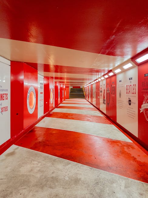 A brightly lit underground pedestrian walkway with red and white painted walls and ceiling. The corridor has a red and white striped floor, with some areas showing signs of wear. On the right side, there are large posters or advertising panels, some showing text related to health and food, mounted on the red walls. The walkway features overhead lighting in a continuous row, illuminating the space evenly. At the far end, a staircase with black handrails leads up to the street level. This environment is typical of transit areas in urban settings, where building access points are connected by a tunnel or underground passage. Such spaces are often used during home relocation or furniture transport to facilitate safe movement between different parts of a building or to prepare items for loading onto vehicles. Man and Van Marylebone may utilize such environments during the moving or packing process, though no furniture or moving equipment is visible in this image.